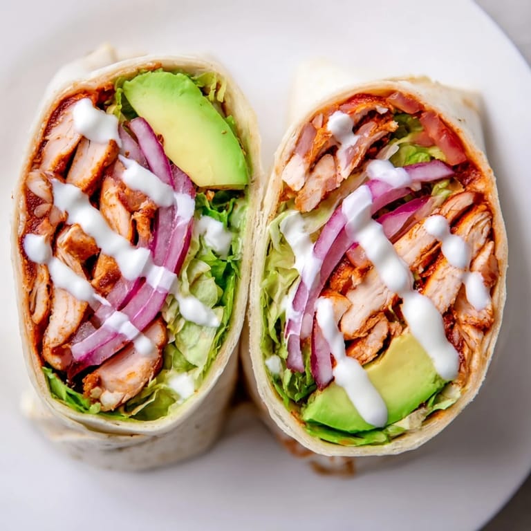 Freshly assembled Smoky Chipotle Chicken Wrap cut in half, revealing vibrant layers of smoky chicken, red onion, tomatoes, and cool avocado with tangy sauce.