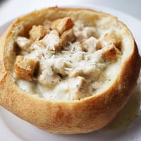 A close-up of a golden bread bowl brimming with creamy Chicken Alfredo, topped with melted Parmesan and fresh parsley.